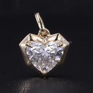 May include: A heart-shaped pendant with a brilliant-cut diamond set in a gold frame. The pendant has a loop at the top for a chain. The background is dark, highlighting the sparkle of the diamond.