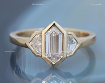 May include: A gold ring featuring a central, elongated, clear gemstone flanked by two smaller, triangular clear gemstones. The ring has a unique geometric design with a polished finish, reflecting light. The ring is set against a blurred, reflective background.
