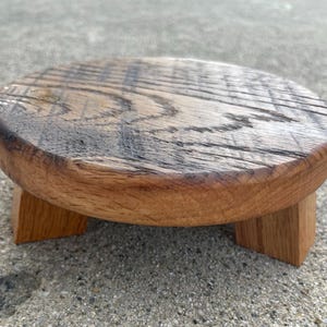 May include: A small, round wooden plant stand with a natural finish. The stand has a circular top and three square legs. The wood grain is visible, and the stand is approximately 6 inches in diameter.