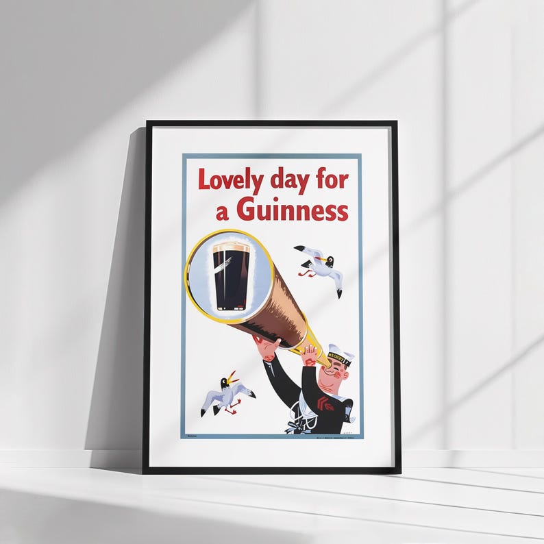 Op de afbeelding: Ingelijste vintage poster met een zeeman die door een telescoop naar een pint Guinness kijkt. De poster heeft de tekst "Lovely day for a Guinness" in het rood. Twee zeemeeuwen vliegen op de achtergrond. De poster zit in een zwarte lijst.