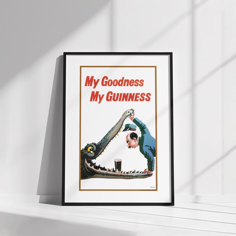 Op de afbeelding: Een ingelijste vintage Guinness-advertentie met een man die een pint stout aan een krokodil aanbiedt. De tekst "My Goodness My Guinness" staat in het rood afgebeeld. De illustratie staat tegen een witte achtergrond.