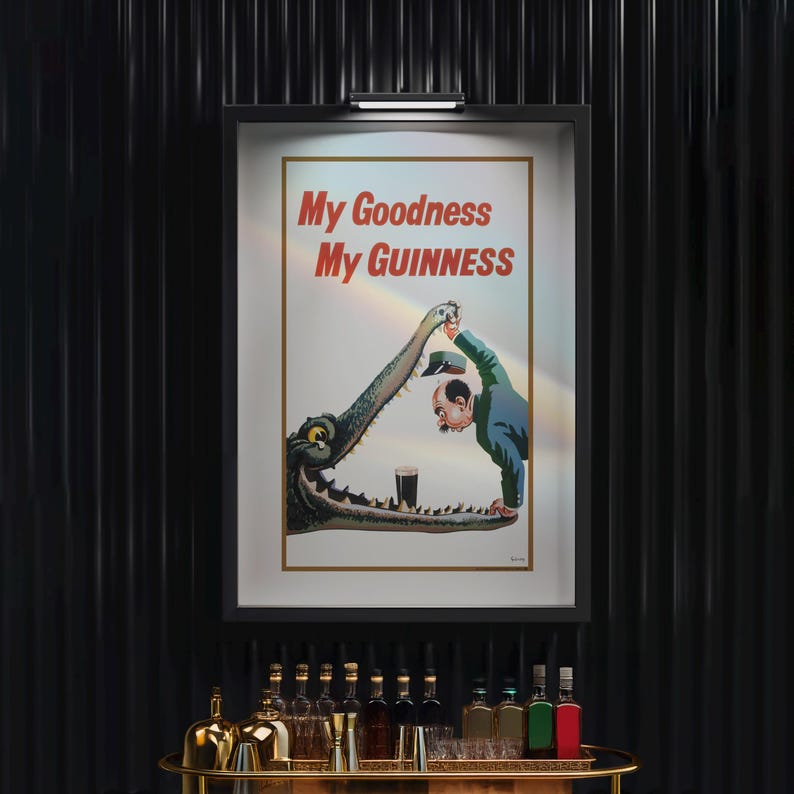 Op de afbeelding: Een ingelijste vintage Guinness-advertentie met de tekst "My Goodness My Guinness". De afbeelding toont een man die een pint Guinness in de bek van een krokodil giet. De poster is zwart omlijst en hangt boven een bar.