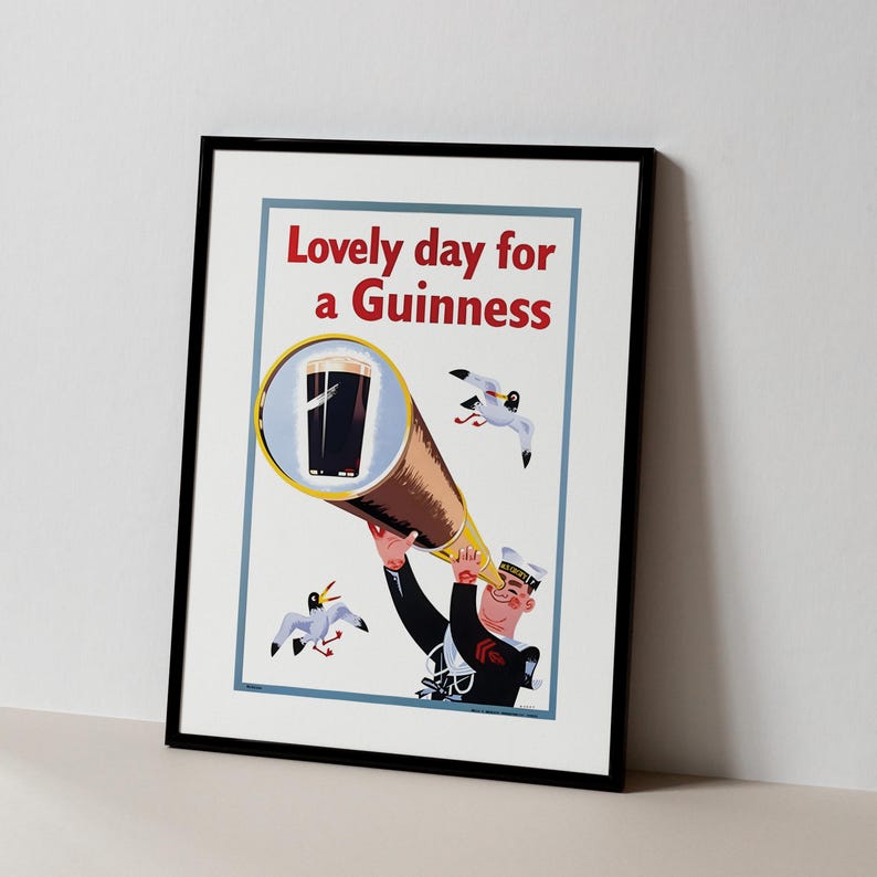 Op de afbeelding: Ingelijste vintage print met een matroos die door een telescoop naar een pint Guinness kijkt. De print heeft de tekst "Lovely day for a Guinness" in het rood. Twee zeemeeuwen vliegen rond de matroos en de pint.