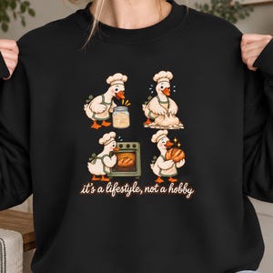 Puede incluir: Una sudadera negra con un gráfico de cuatro patos blancos con gorros de chef y delantales verdes, mostrados horneando pan en varias etapas. Debajo de las ilustraciones, el texto "it's a lifestyle, not a hobby" aparece en letra cursiva blanca.
