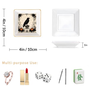 May include: Two square white ceramic trinket trays measuring 4 inches by 4 inches by 1 inch. One tray features a black raven, pumpkins, and floral scrollwork. The other is plain white. Below are a ring, lipstick, two dice, pens, and a mahjong tile.