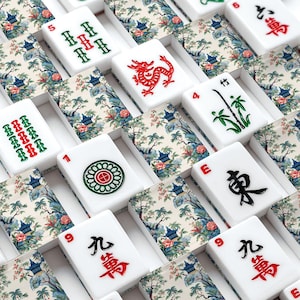 Op de afbeelding: Een verzameling witte mahjongstenen met een decoratief bloemen- en landschapspatroon op de achterkant. De voorkanten tonen diverse gegraveerde symbolen in rood, groen en zwart, waaronder Chinese karakters, bamboestengels, draken en cirkels.