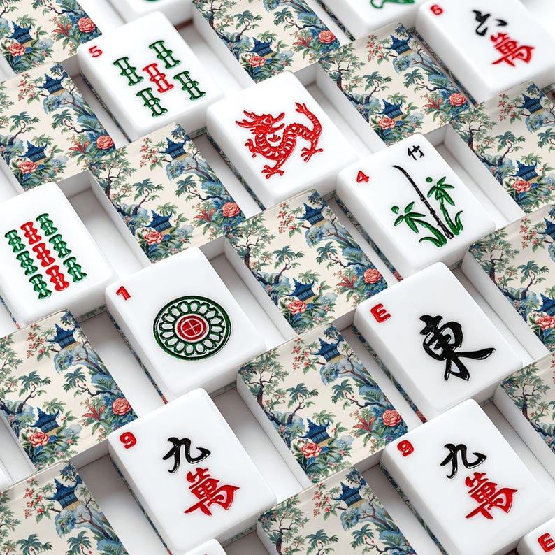 Op de afbeelding: Een verzameling witte mahjongstenen met een decoratief bloemen- en landschapspatroon op de achterkant. De voorkanten tonen diverse gegraveerde symbolen in rood, groen en zwart, waaronder Chinese karakters, bamboestengels, draken en cirkels.