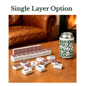 May include: A set of white mahjong tiles with colorful patterned backs and red or black character faces, arranged in a single layer. Next to the tiles is a white beverage can cooler featuring the green text MAHJONG and tile illustrations.
