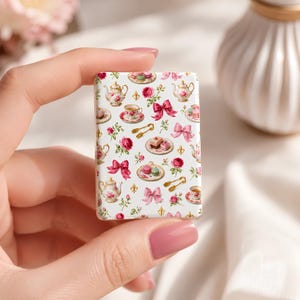 May include: A rectangular magnet featuring a white background with a repeating pattern of floral tea sets, pink bows, gold sugar tongs, and small plates with macarons and cakes. The design includes detailed illustrations of roses and teapots.