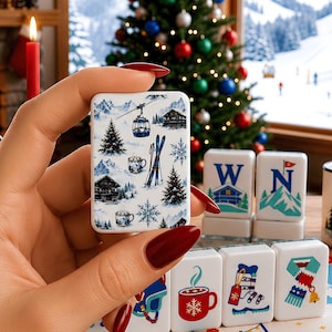 May include: A rectangular white tile featuring a winter scene with blue mountains, a cable car, evergreen trees, skis, snowflakes, and mugs of hot cocoa. Other tiles nearby display letters W and N, a red mug, a ski boot, and a scarf.
