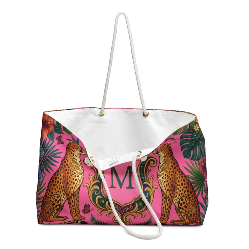 May include: A pink tote bag with a white interior and rope handles. The bag features a design of two cheetahs, tropical flowers, and foliage. A gold-framed initial "M" is centered on the bag.