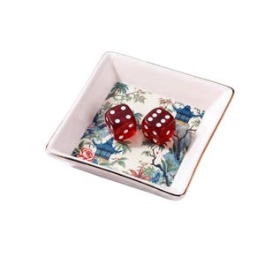 May include: A square, white ceramic dish with a gold rim, containing two red dice with white dots. The dish's interior features a floral and landscape pattern in blue, green, and pink.