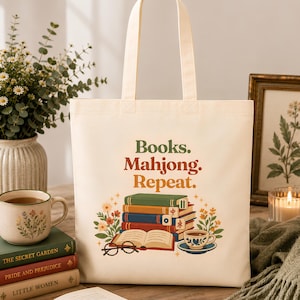 Op de afbeelding: Een crèmekleurige canvas tas met de tekst "Books. Mahjong. Repeat." in groen, rood en oranje. De afbeelding toont een stapel kleurrijke boeken met mahjongstenen, een open boek, een bril, een theekopje en bloemmotieven als decoratie.