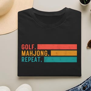 May include: Black t-shirt with the words "GOLF. MAHJONG. REPEAT." in a retro design. The text is in red, gold, and teal horizontal stripes. A casual tee for golf and mahjong enthusiasts.