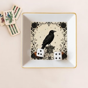 May include: A square white ceramic tray with a gold rim featuring a central illustration of a black raven perched on a pedestal, surrounded by a floral border with roses, stars, and a skull. Two white six-sided dice with black pips rest inside.