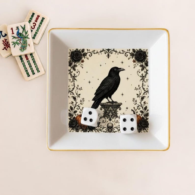May include: A square white ceramic tray with a gold rim featuring a central illustration of a black raven perched on a pedestal, surrounded by a floral border with roses, stars, and a skull. Two white six-sided dice with black pips rest inside.