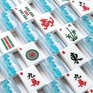May include: A close-up shot of a Mahjong set. The white tiles feature various symbols and numbers in red, green, and black. The set includes tiles with a red dragon, bamboo, and floral designs. The background features a blue and white wave pattern.