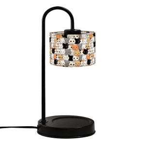 May include: A black desk lamp with a curved arm and a round base. The lampshade is cylindrical and covered in a repeating pattern of cartoon cats in various colors. The lamp is plugged into an electrical outlet.