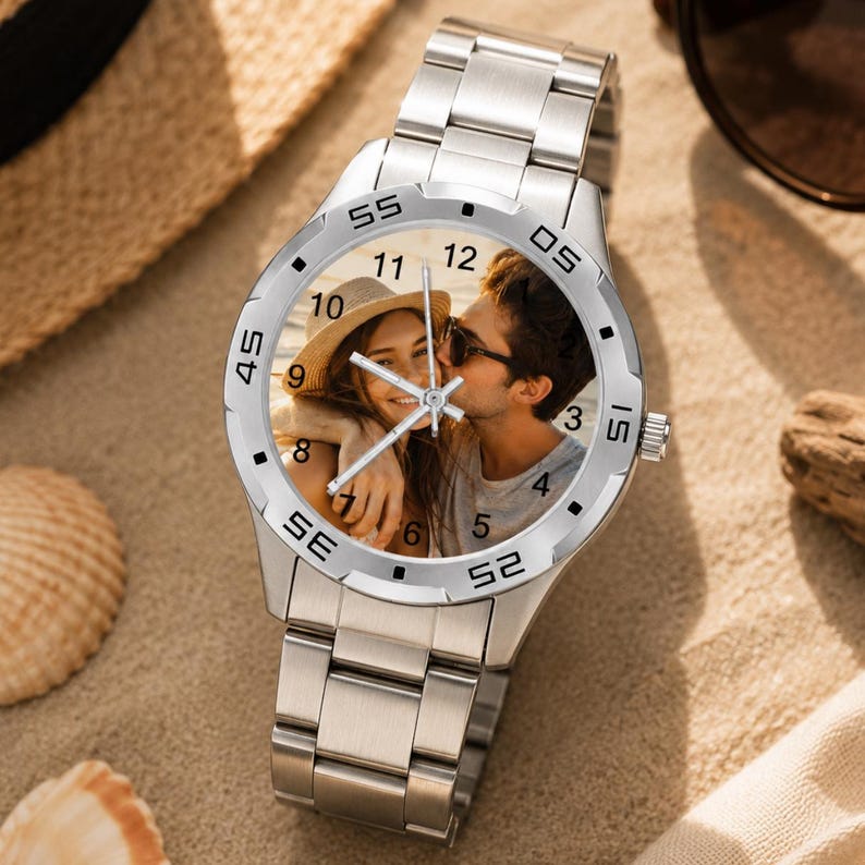 May include: A silver wristwatch with a photo of a couple kissing on the watch face. The watch has a stainless steel band and a round face with black numerals. The watch is on a sandy surface.