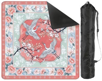 May include: A square yoga mat with a floral and crane design in shades of pink, blue, and white. The mat has a black underside and comes with a black carrying bag with a shoulder strap. The mat features a decorative border.