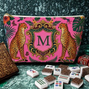 May include: A pink zippered cosmetic bag with a cheetah print design. The bag features two cheetahs flanking a shield with the letter "M". Tropical foliage and a gold zipper complete the design. Mahjong tiles are scattered in the foreground.