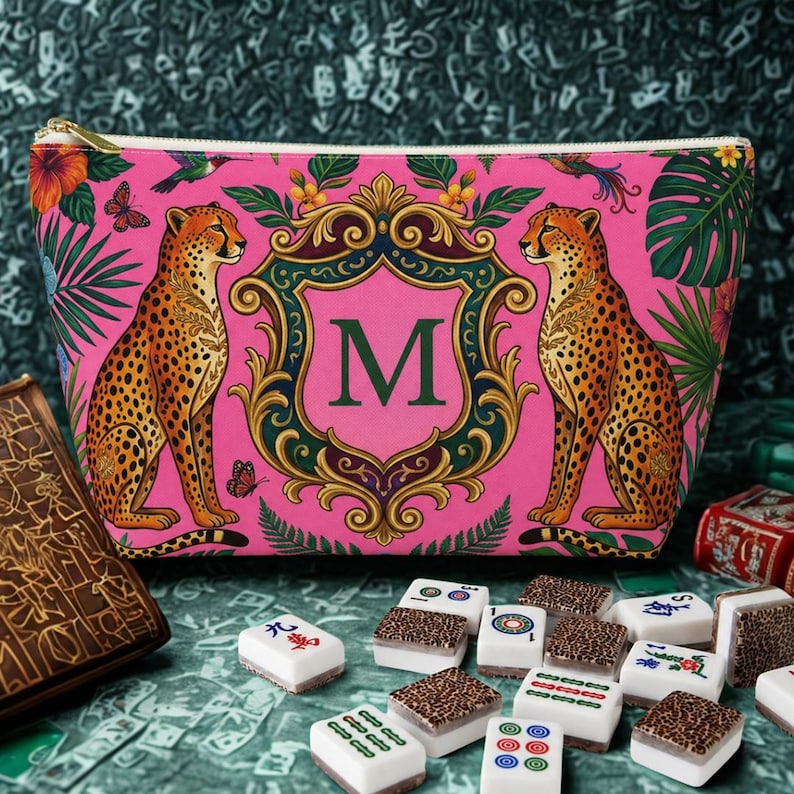 May include: A pink zippered cosmetic bag with a cheetah print design. The bag features two cheetahs flanking a shield with the letter "M". Tropical foliage and a gold zipper complete the design. Mahjong tiles are scattered in the foreground.