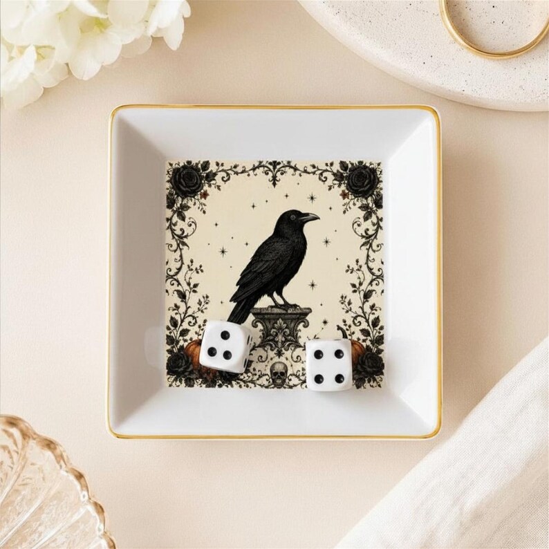 May include: A square white ceramic trinket dish with a gold-toned rim. The center features a black raven illustration perched on a pedestal, surrounded by a decorative floral border, small stars, and a skull. Two white dice rest inside the dish.