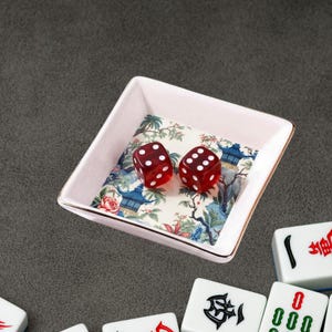May include: A square, light pink dish with a gold rim, holding two red dice. The dish's interior features a colorful Asian-inspired floral and architectural design. Mahjong tiles are visible in the foreground, suggesting a game theme.