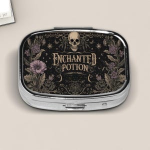 May include: A silver pill box with a black background featuring the words "Enchanted Potion" in a vintage font, a skull, and floral accents in purple and beige. The pill box is rectangular with rounded edges.