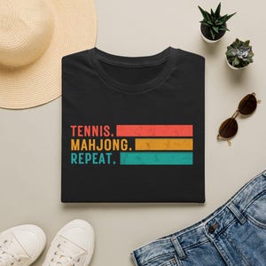 May include: Black t-shirt with the text "TENNIS. MAHJONG. REPEAT." in a retro font. The text is layered over three horizontal stripes in red, gold, and teal. The shirt is displayed with a straw hat, sunglasses, and denim jeans.