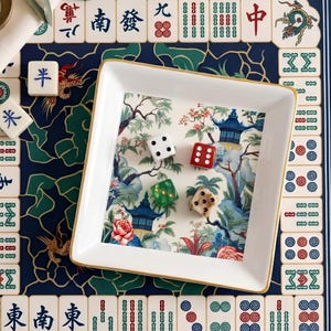 May include: A white square tray with a gold rim, containing four dice. The tray has a scenic Asian-inspired design with pagodas and floral elements. The tray is set on a blue patterned surface with Mahjong tiles.