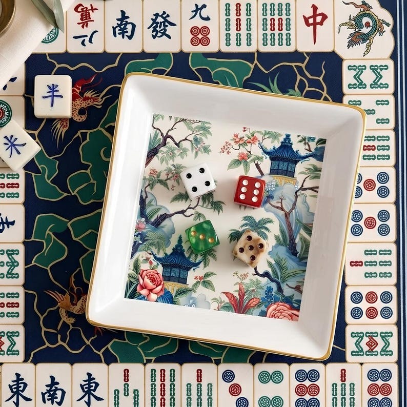 May include: A white square tray with a gold rim, containing four dice. The tray has a scenic Asian-inspired design with pagodas and floral elements. The tray is set on a blue patterned surface with Mahjong tiles.