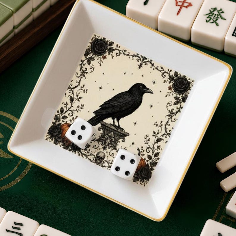 May include: A square white ceramic tray with a gold-trimmed edge featuring a central illustration of a black crow perched on a pedestal, surrounded by roses, vines, and a small skull. Two white six-sided dice with black pips rest inside the tray.