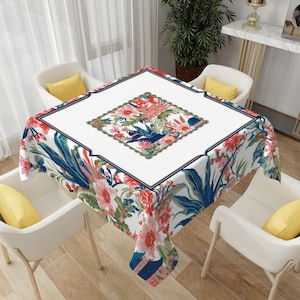 May include: A square tablecloth with a white background and a floral pattern. The pattern includes red, pink, and blue flowers and green leaves. The tablecloth has a navy blue border and is set on a table with white chairs.