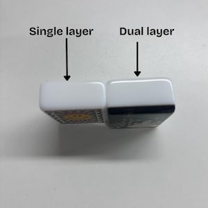 Puede incluir: Dos fichas rectangulares con lados blancos y reverso negro, colocadas una al lado de la otra. La ficha izquierda est&aacute; etiquetada como Single layer con una flecha, y la derecha como Dual layer con una flecha. La ficha izquierda tiene un sol amarillo.