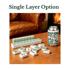 May include: A set of white mahjong tiles with green marbled backs and red or black character designs, arranged in a single layer. A white beverage can cooler with green text reading MAHJONG and small tile illustrations sits next to the tiles.
