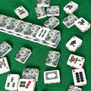 Op de afbeelding: Een verzameling rechthoekige mahjongtegels met witte oppervlakken met zwarte, groene en rode inkttekens, naast tegels versierd met een blauw en groen landschapspatroon van bomen, pagodes en bloemen, verspreid over een groen oppervlak.