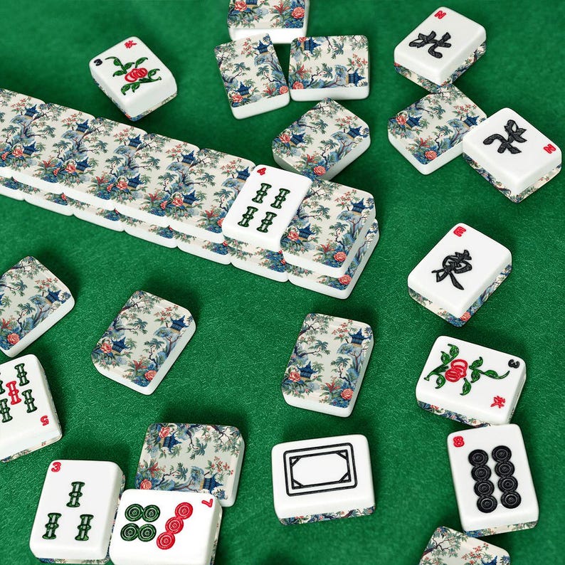 Op de afbeelding: Een verzameling rechthoekige mahjongtegels met witte oppervlakken met zwarte, groene en rode inkttekens, naast tegels versierd met een blauw en groen landschapspatroon van bomen, pagodes en bloemen, verspreid over een groen oppervlak.