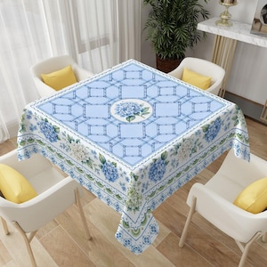 May include: A square tablecloth featuring a light blue geometric lattice pattern with a central circular medallion containing blue hydrangea flowers. The border displays a repeating floral motif with blue and white hydrangea clusters and green leaves.