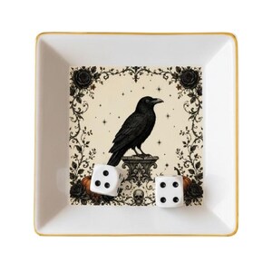 May include: A square white ceramic tray with a gold-toned rim featuring a central illustration of a black raven perched on a pedestal, surrounded by black roses, vines, small stars, and a skull. Two white six-sided dice rest inside the tray.
