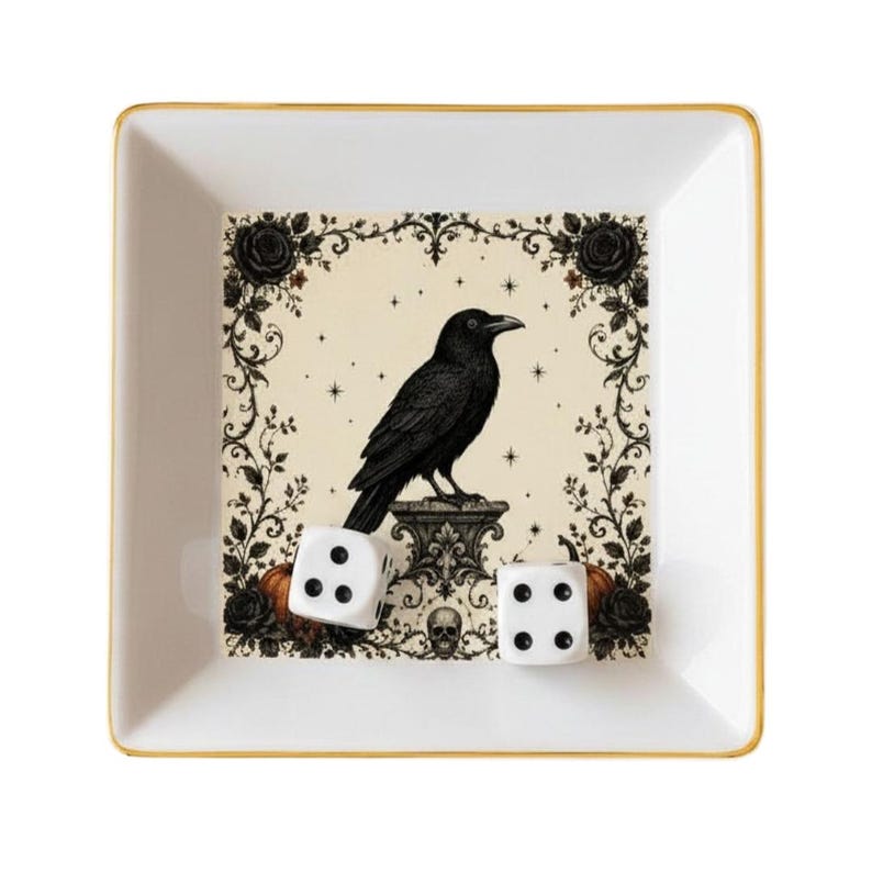 May include: A square white ceramic tray with a gold-toned rim featuring a central illustration of a black raven perched on a pedestal, surrounded by black roses, vines, small stars, and a skull. Two white six-sided dice rest inside the tray.