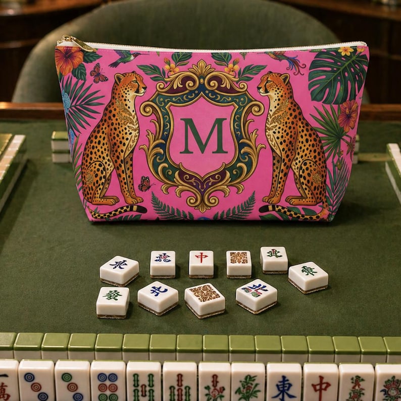 May include: A pink zippered cosmetic bag featuring two cheetahs flanking a shield with the letter "M". The bag is decorated with tropical foliage and butterflies. Mahjong tiles are in the foreground.