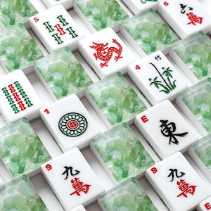 May include: A close-up shot of a Mahjong set with white tiles featuring red, green, and black symbols and characters. The set includes tiles with dragons, bamboo, and other traditional designs. The tiles are arranged in a grid.