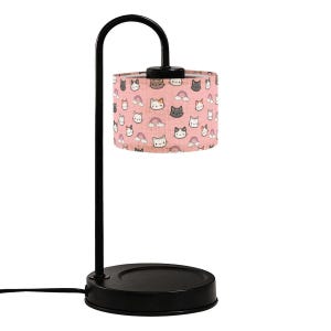 May include: Black metal desk lamp with a curved arm and a round base. The lampshade is cylindrical and features a pink background with a repeating pattern of cat faces and rainbows. The lamp is plugged into an electrical outlet.