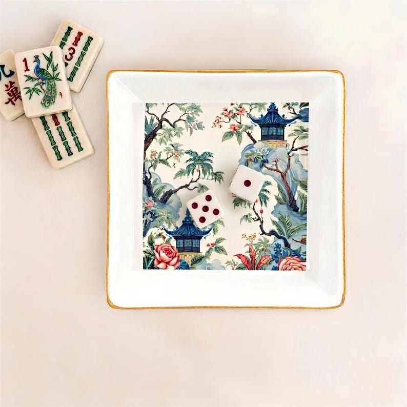 May include: A white square trinket dish with a gold rim, featuring a scenic Asian-inspired design with pagodas, flowers, and trees. Two white dice with red dots and several Mahjong tiles are next to the dish. The Mahjong tiles have green and red markings.