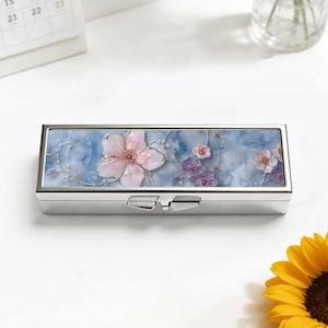May include: A rectangular, silver-colored pill box with a floral design. The lid features a light blue background with pink and purple flowers. The box has a clasp closure and a shiny finish.