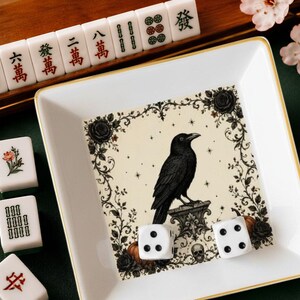 May include: A square white ceramic tray with a gold rim featuring a central illustration of a black crow perched on a stone pedestal, surrounded by black roses, vines, and a small skull. Two white six-sided dice rest inside the tray.