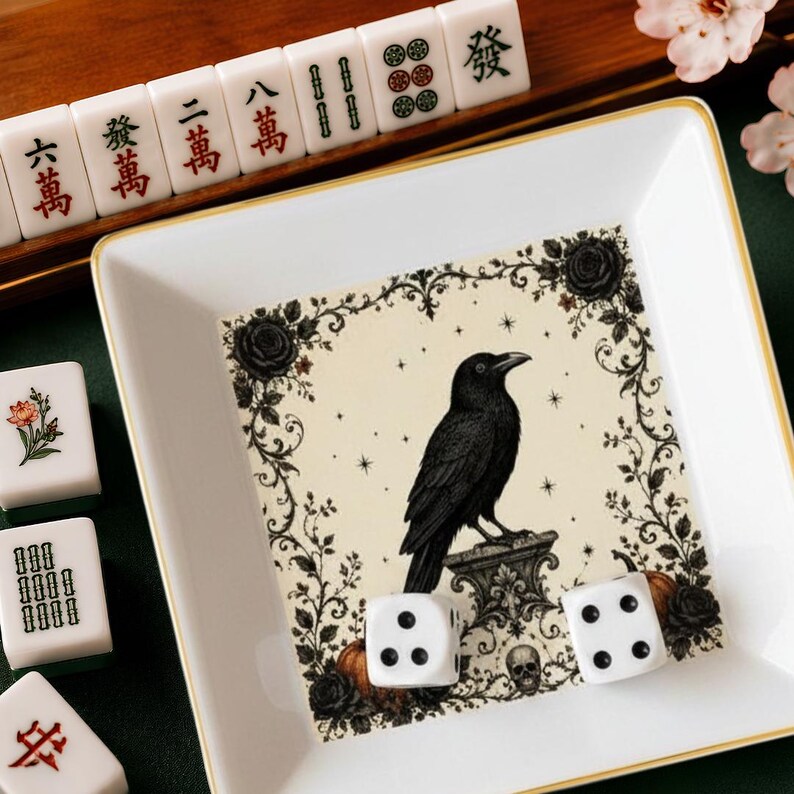 May include: A square white ceramic tray with a gold rim featuring a central illustration of a black crow perched on a stone pedestal, surrounded by black roses, vines, and a small skull. Two white six-sided dice rest inside the tray.