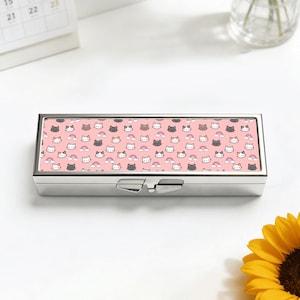 May include: A rectangular, silver-colored pill box with a hinged lid. The top surface features a repeating pattern of cartoon cat faces, rainbows, and clouds on a pink background. The box is closed and sits on a white surface.