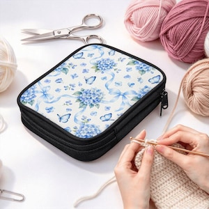 May include: A rectangular black zippered case featuring a white surface with a blue floral and butterfly pattern. The case is surrounded by balls of yarn, a pair of silver scissors, and a metal crochet hook working on a beige yarn project.